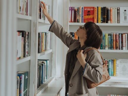 A young woman reaching for a book on a stylish library bookshelf, embodying education and leisure.